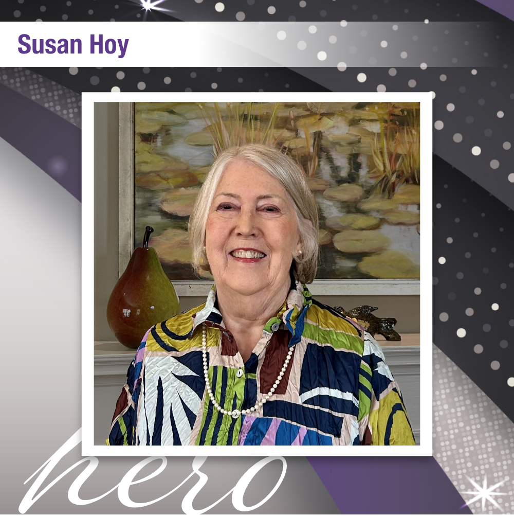 Photo of Susan Hoy that reads "hero". Susan is posing in front of a painting, smiling and wearing a colorful dress.